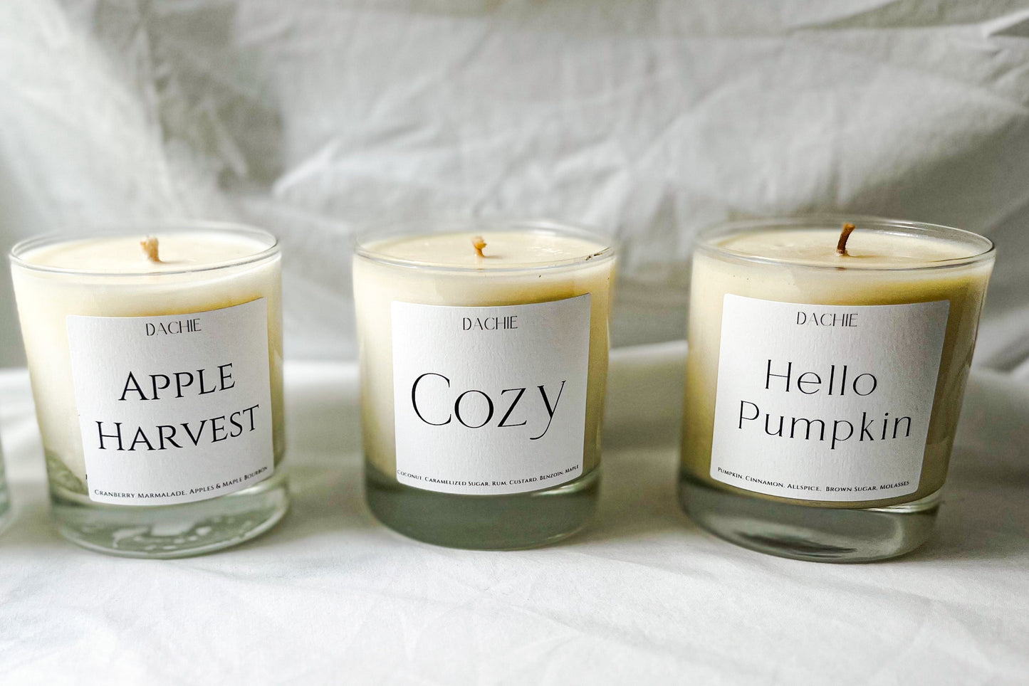 Four candles with labels 'Autumn', 'Apple Harvest', 'Cozy', and 'Hello Pumpkin' on a white fabric background.